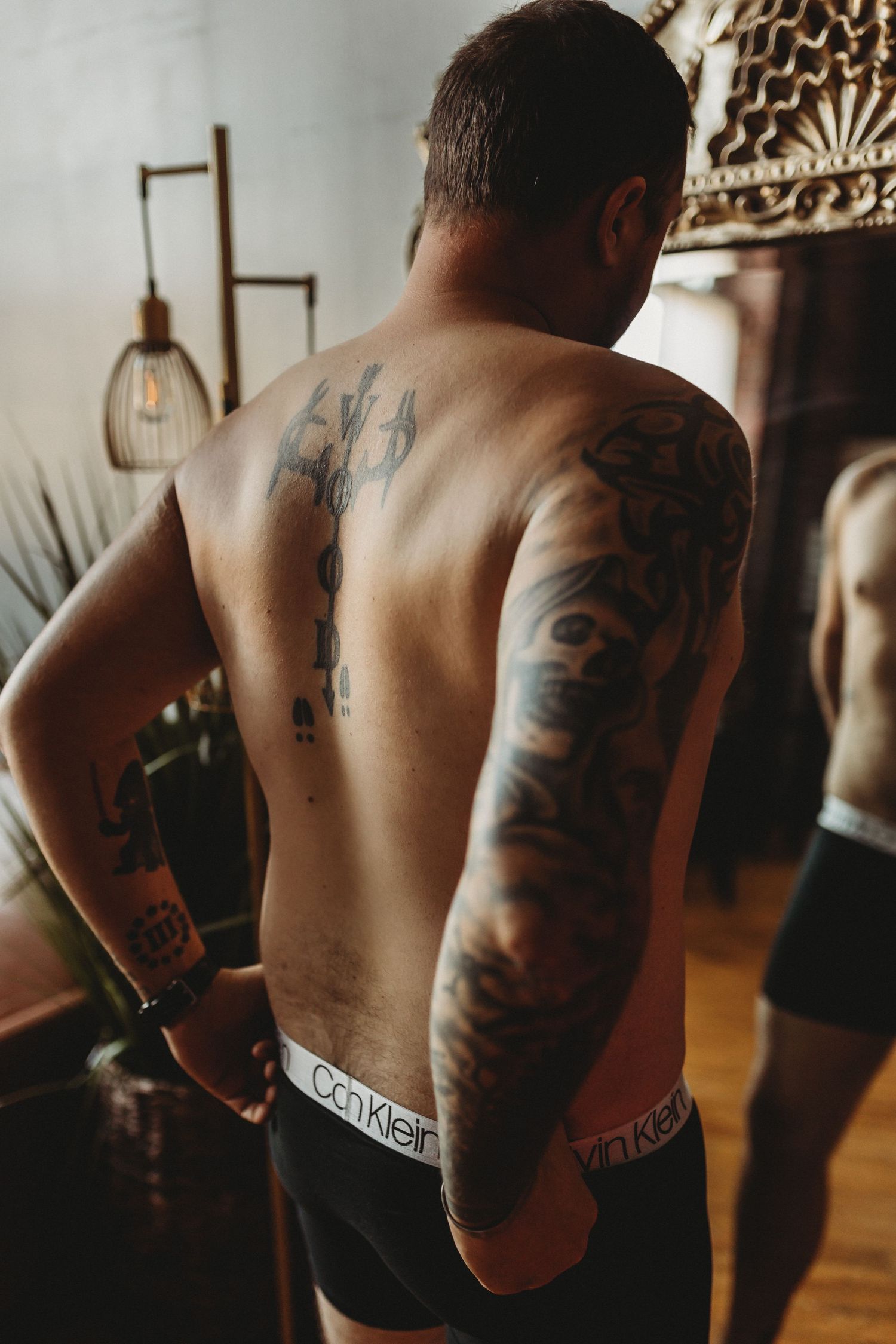 Back view showing detailed sleeve tattoos and dark underwear against indoor lighting.