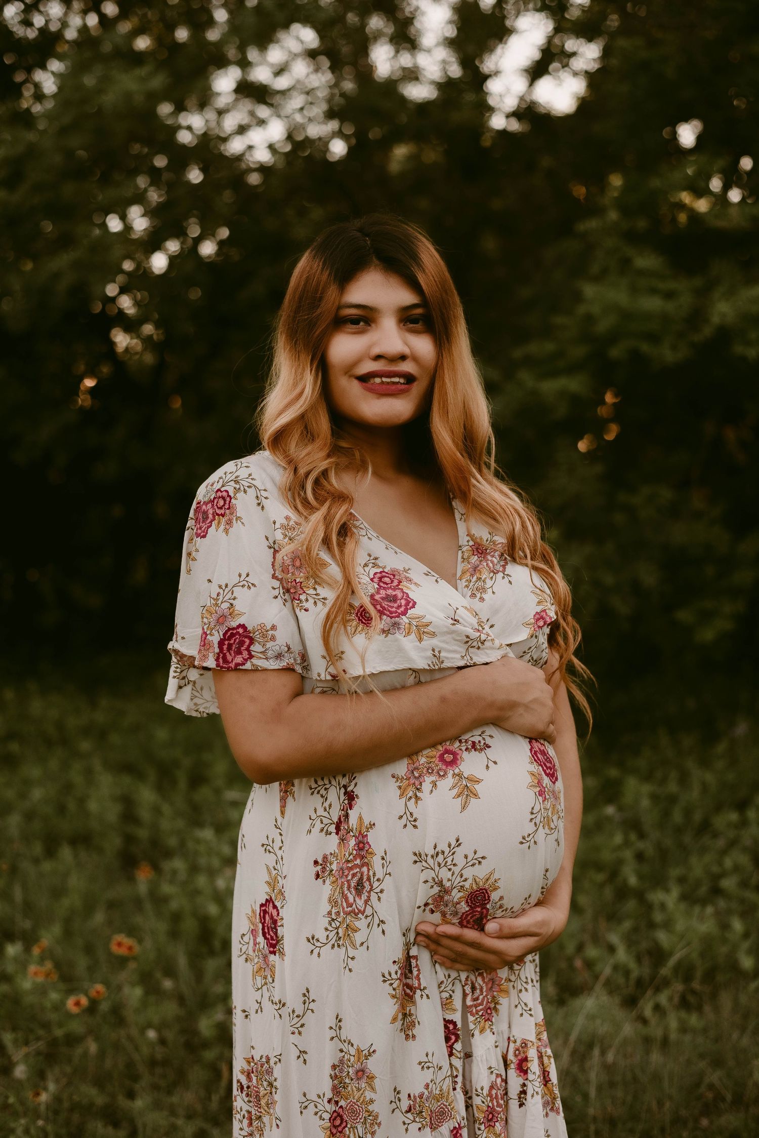 Expectant mother wearing white floral dress poses in grassy field during sunset photoshoot.