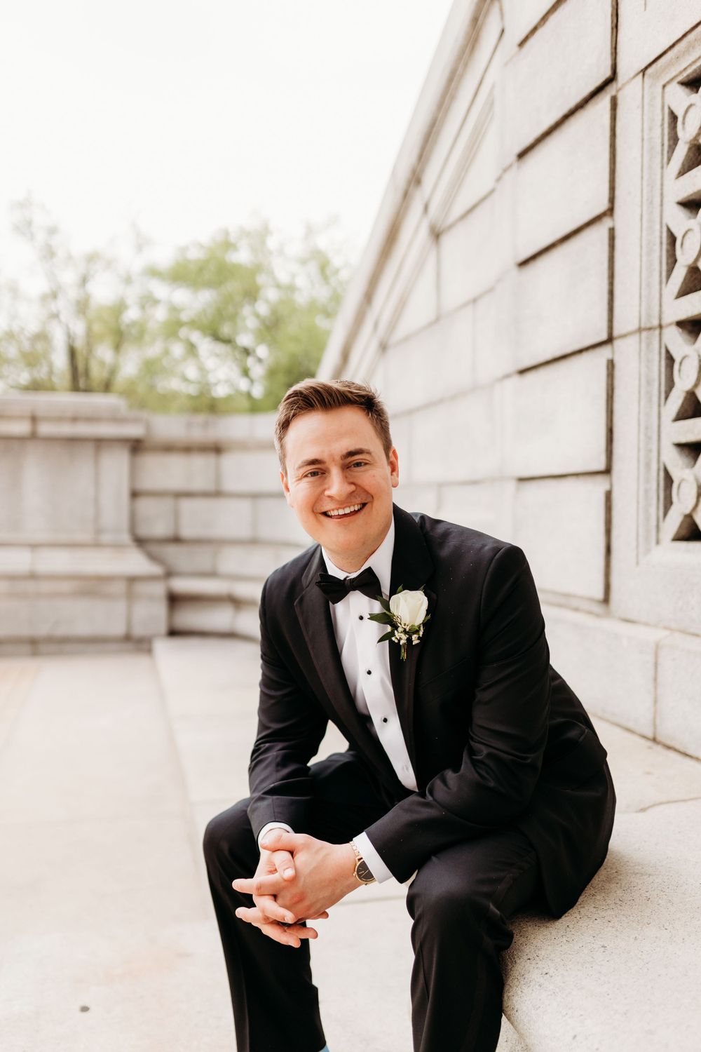 Groom in classic black tuxedo sits on stone steps during wedding portraits.