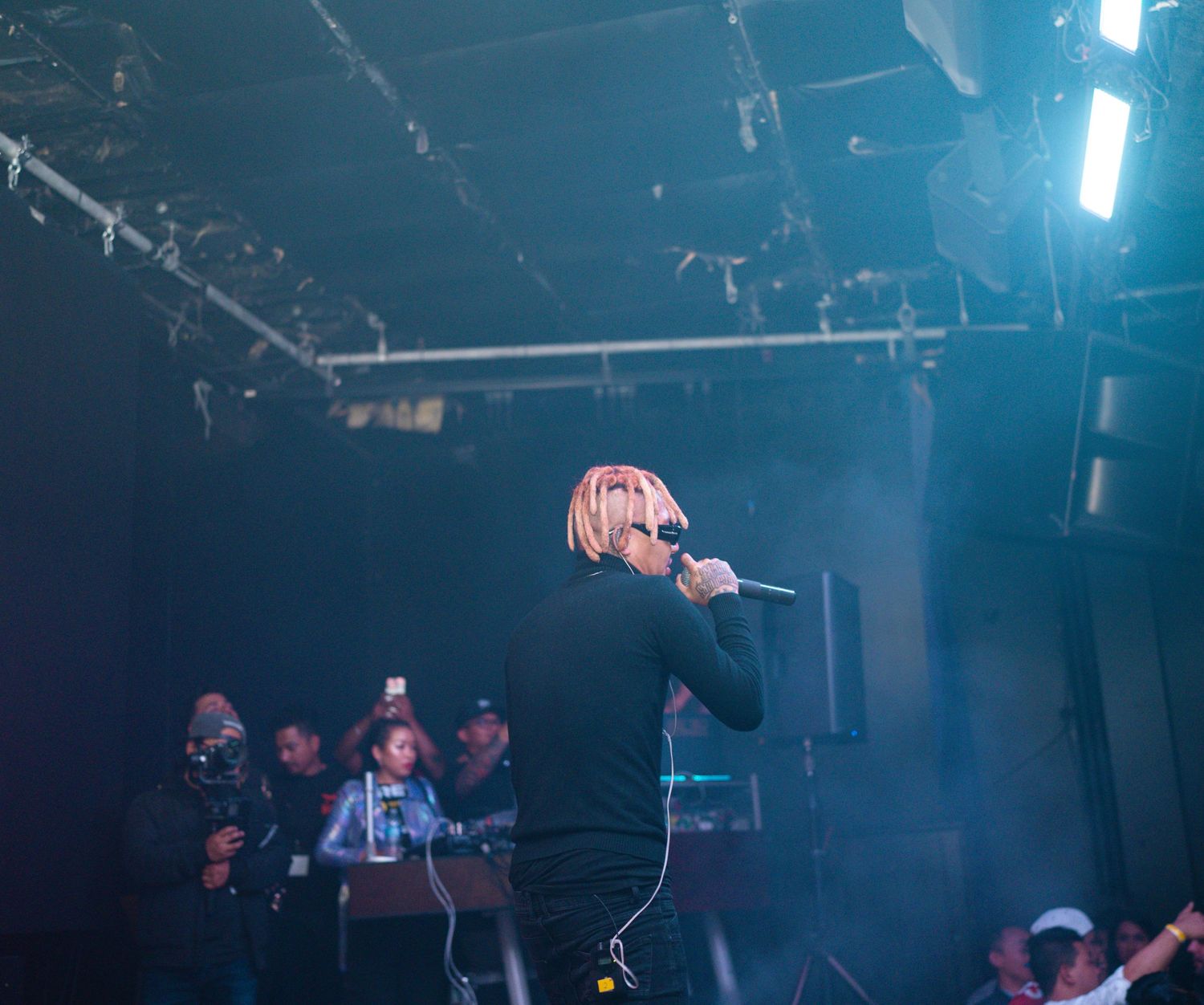 Concert performer in black outfit on stage with dramatic blue lighting and crowd in background.