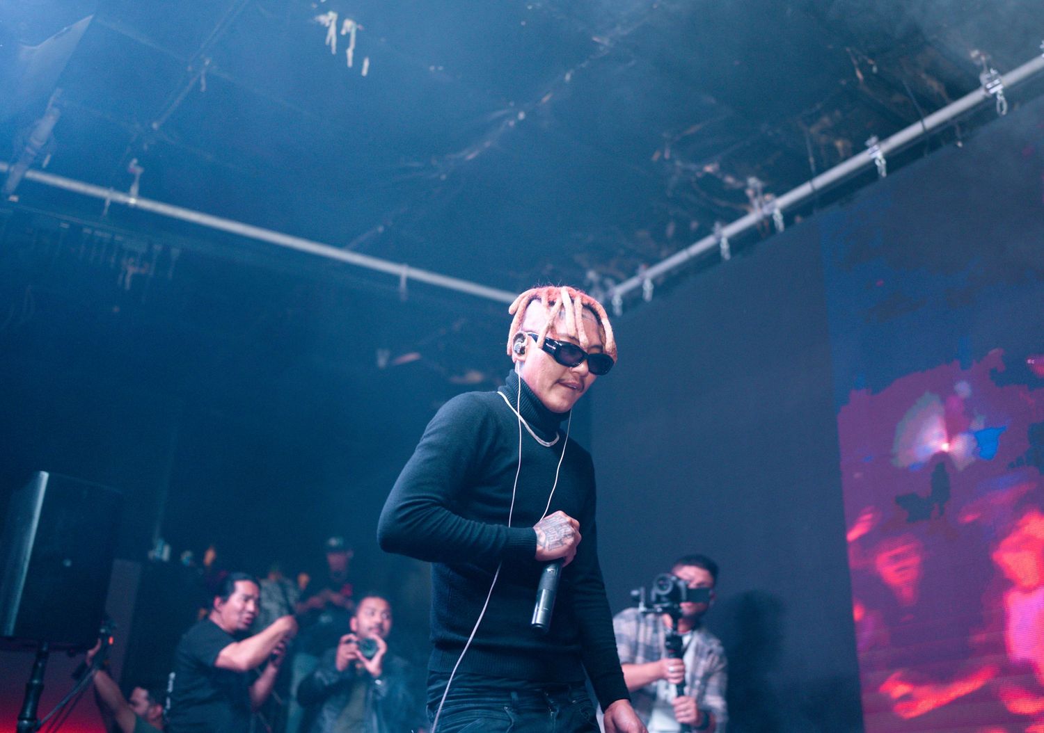 Performer with light hair in black attire singing on dimly lit concert stage.