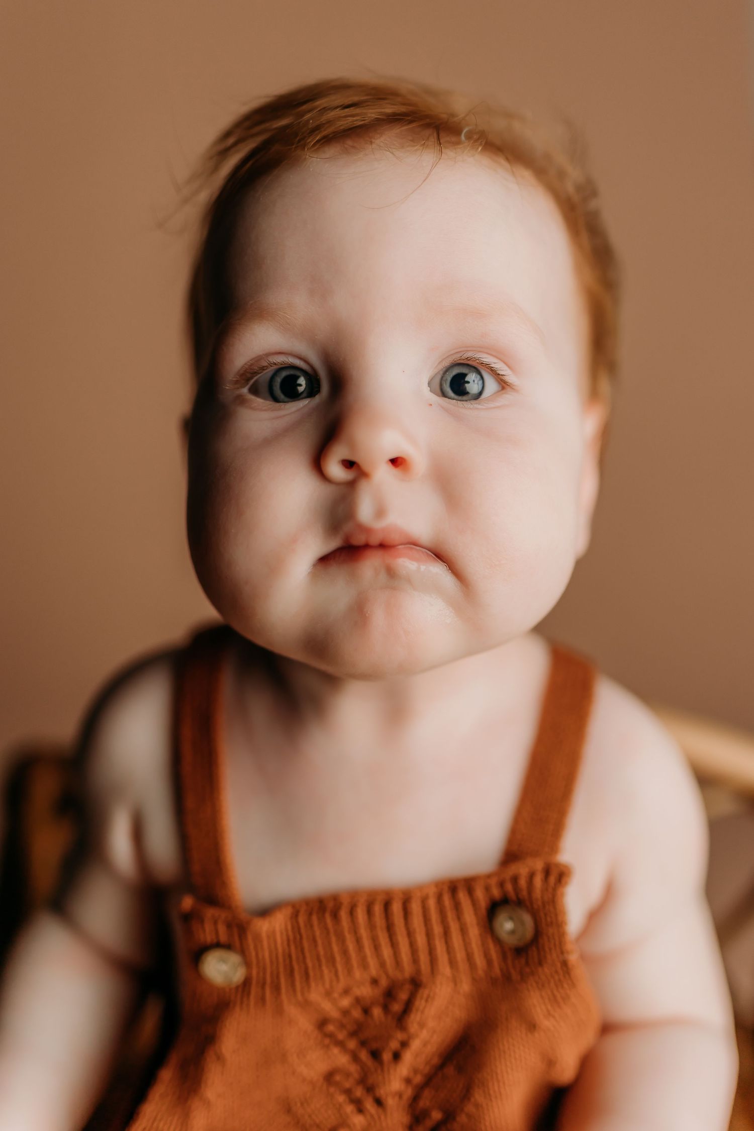 Close up portrait of baby wearing orange overalls against neutral background.