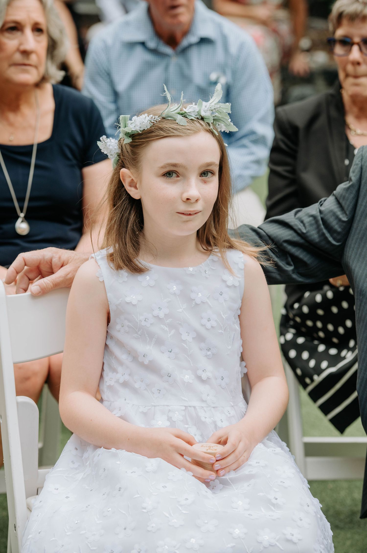 Young attendant in white dress and floral crown seated during outdoor ceremony.