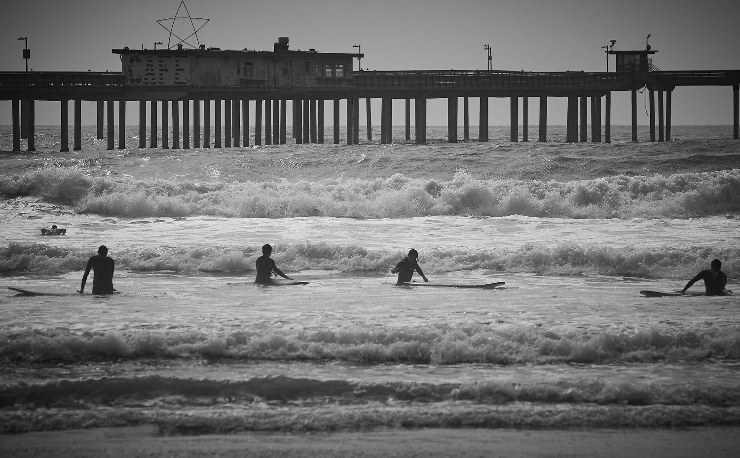Black and white photo of surfers catching waves near a coastal pier.