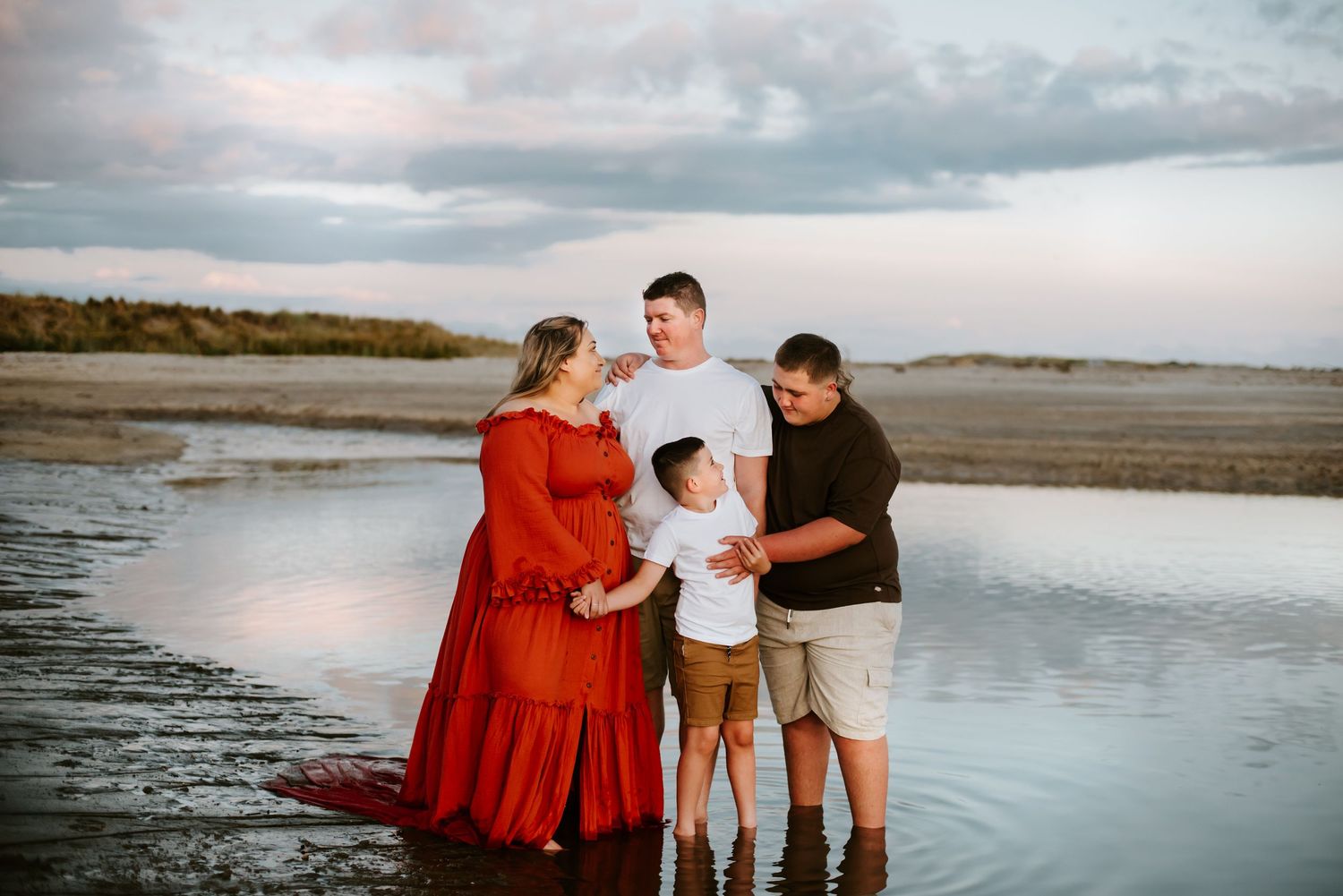 A family of four stands together at the beach during sunset, with the mother wearing a flowing red dress.