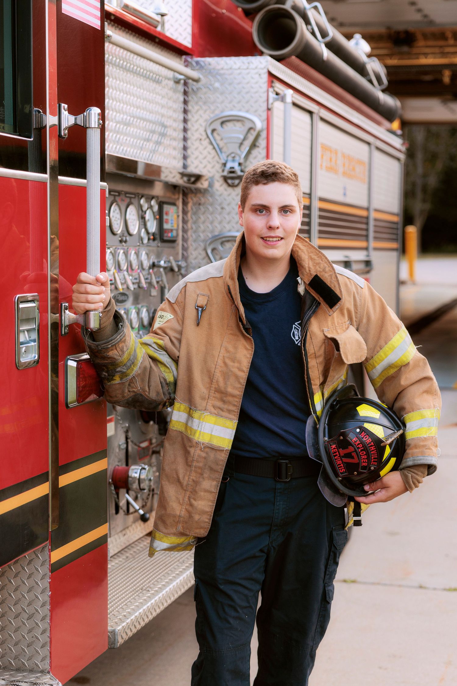 A firefighter in turnout gear holds their helmet while posing next to their fire engine.