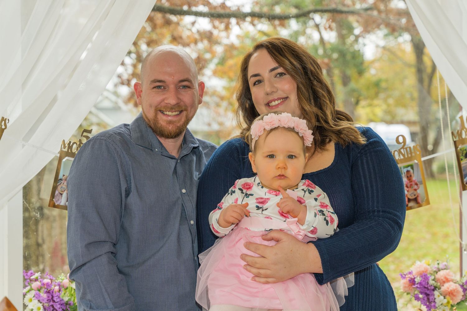 Happy family portrait with man, woman, and baby girl in floral outfit at outdoor autumn event.