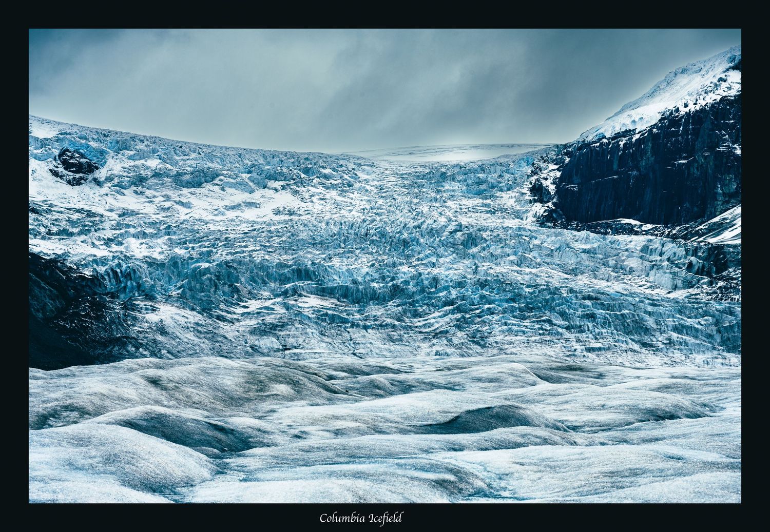 Massive blue glacier field stretches across snow-covered mountain terrain under moody sky.