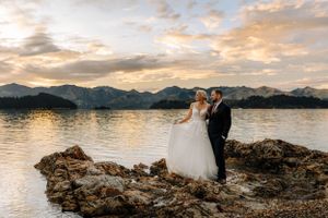 bride and groom standing on rocks looking out across Christchurch harbour at sunset with port hills in background