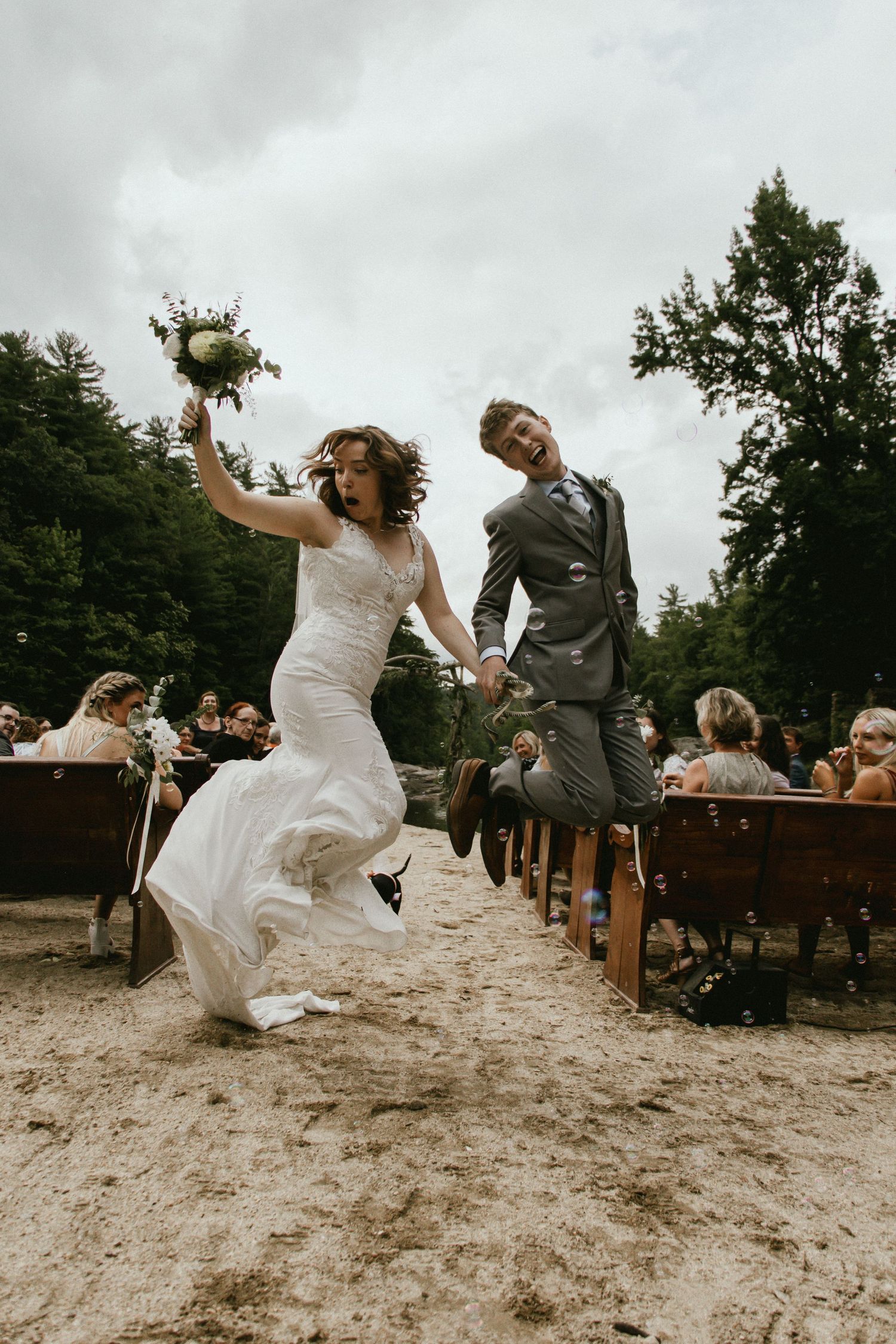 Newlyweds joyfully jumping and celebrating while walking down the aisle at outdoor ceremony.