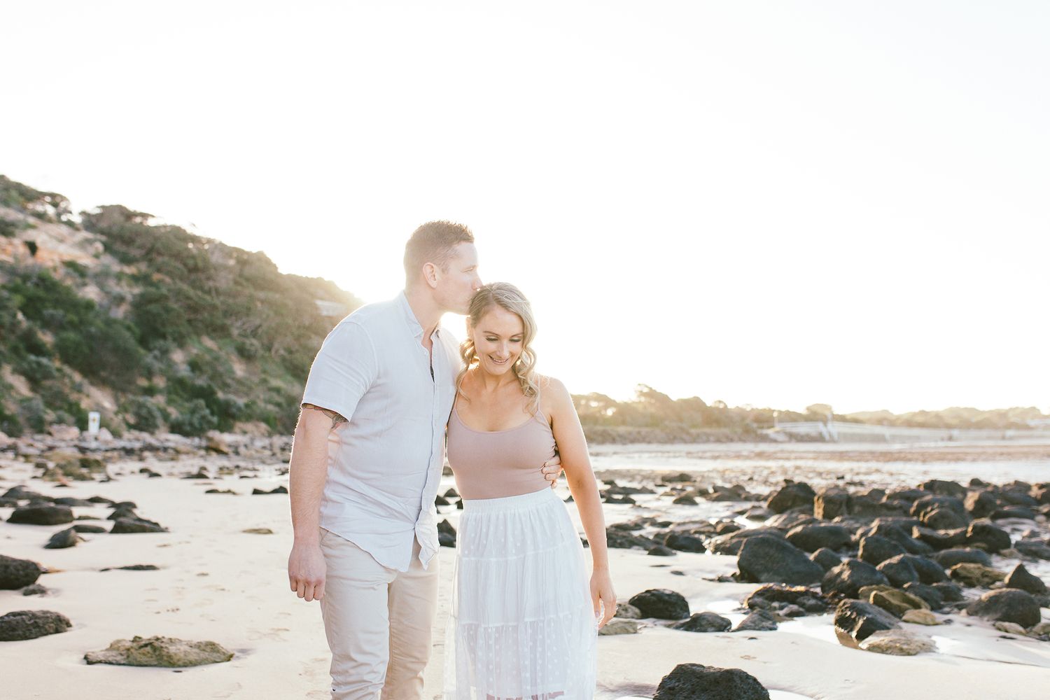 A couple shares a tender moment while walking along a rocky coastline at sunset.