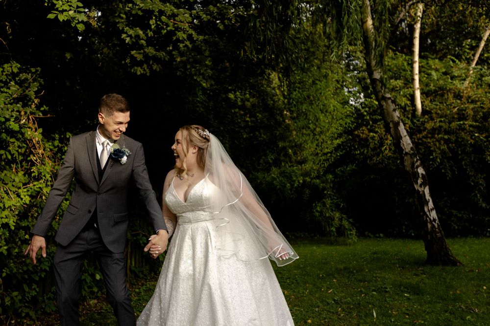 Newlyweds walking hand in hand through a sunlit garden wearing formal wedding attire.