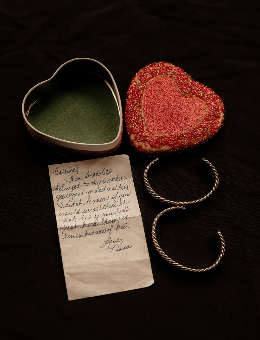 Vintage heart-shaped red jewelry box with silver bracelets and handwritten note on dark background.