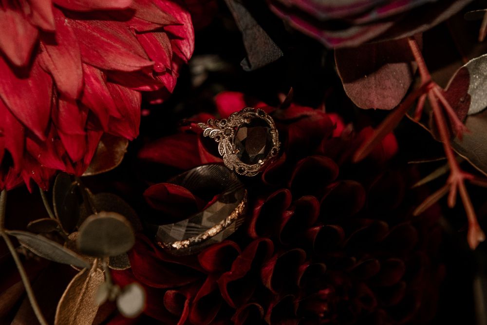 Ornate wedding rings resting on deep red and burgundy floral bouquet in dark moody setting.