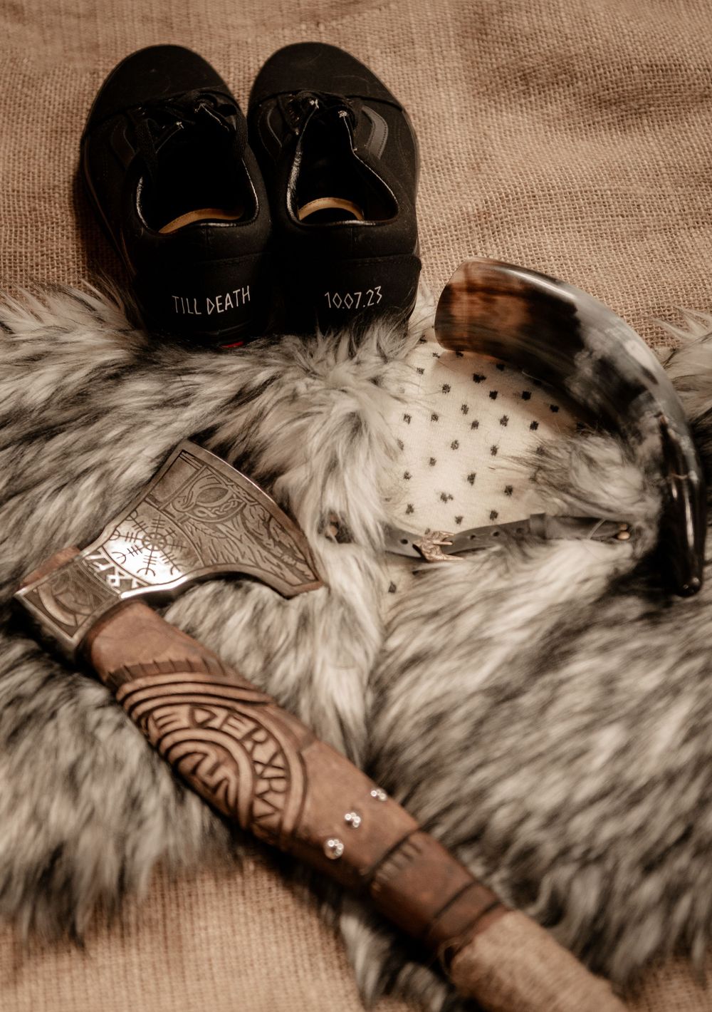 Viking wedding accessories including an ornate axe, drinking horn, and black shoes reading Till Death on a fur pelt.