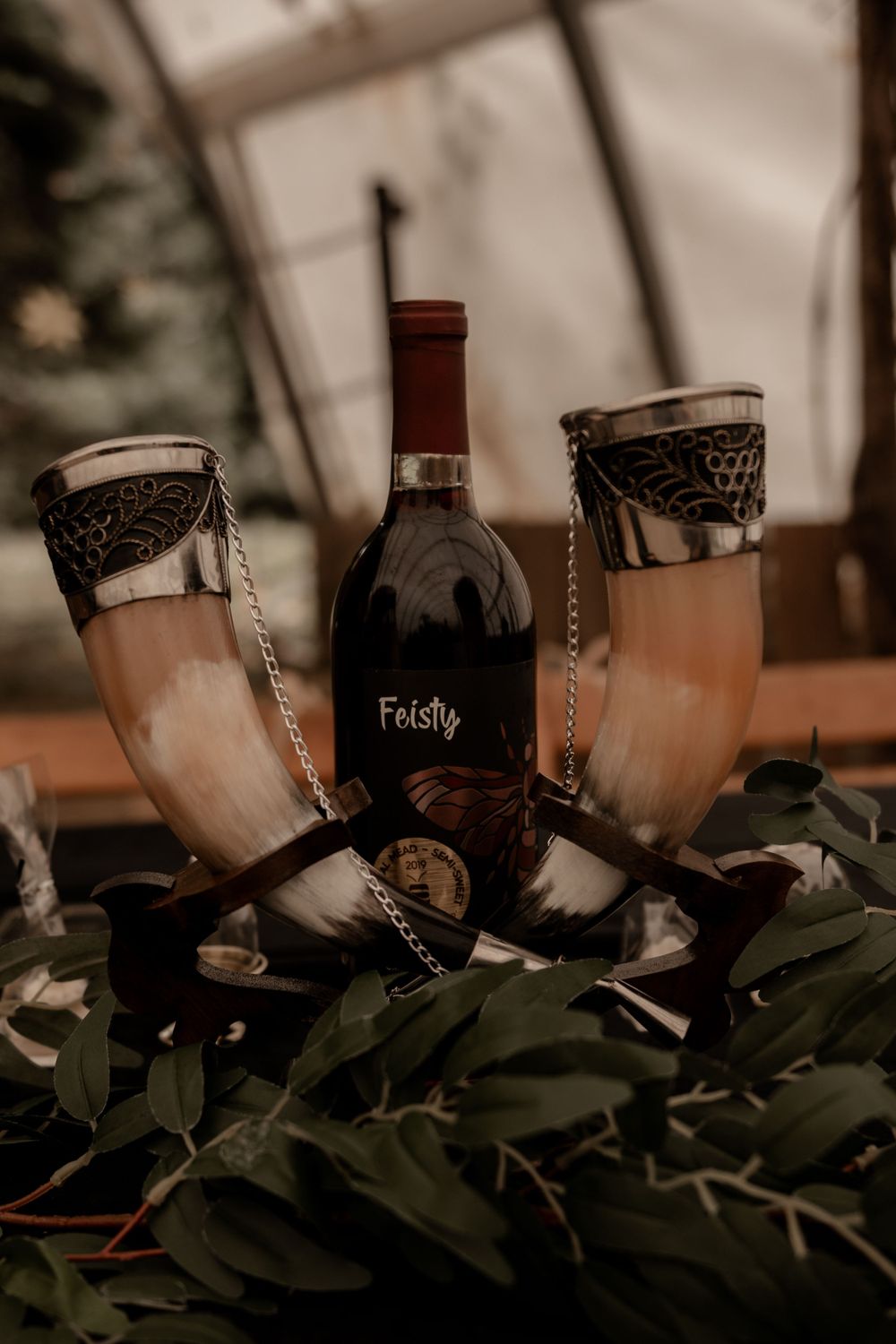Two decorated Viking drinking horns flank a bottle of Feisty red wine on a rustic woodland wedding table.