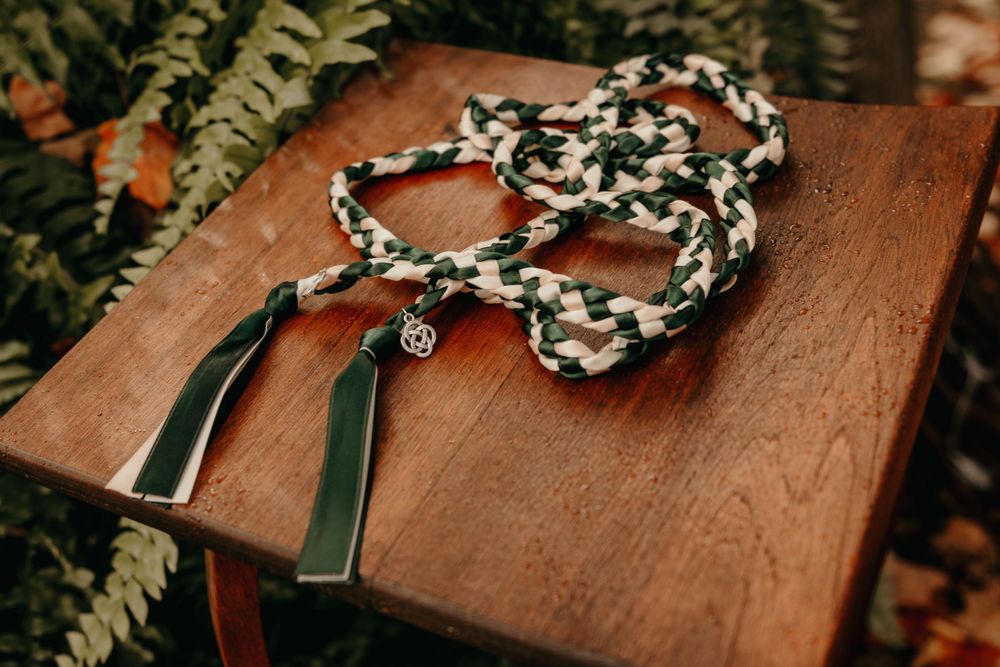 Green and white braided handfasting cord with charm and tassels displayed on a wooden table outdoors.
