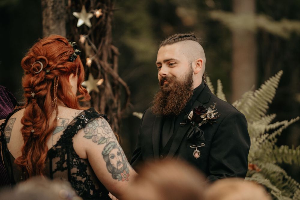 Tattooed bride in black lace dress and groom with beard exchange vows in a forest wedding ceremony.