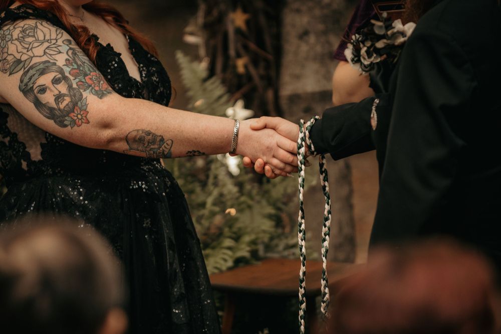 Tattooed bride and groom hold hands during a chain handfasting ceremony at their gothic woodland wedding.