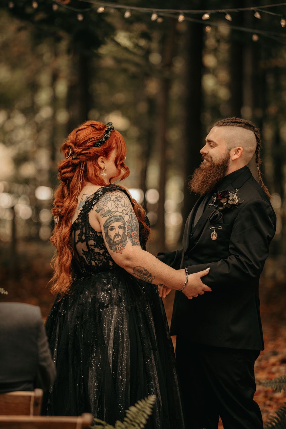 Tattooed redhead bride in black gown and bearded groom hold hands during an outdoor forest wedding ceremony.