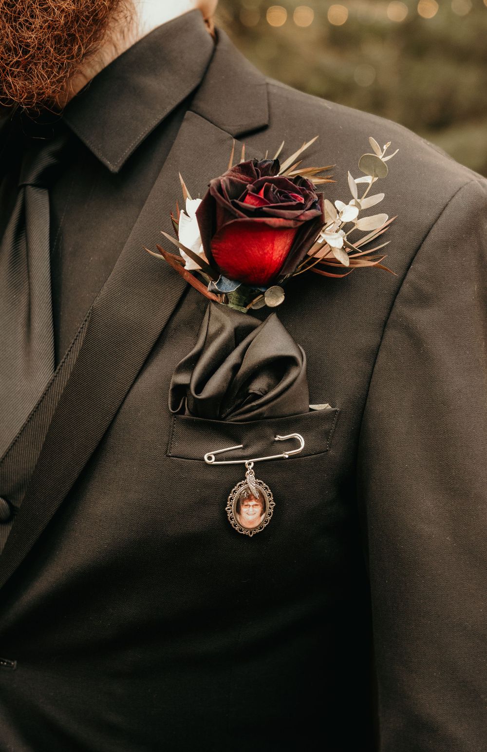 Dark gothic boutonniere with deep red rose, thistle, and memorial locket pin on black suit lapel.