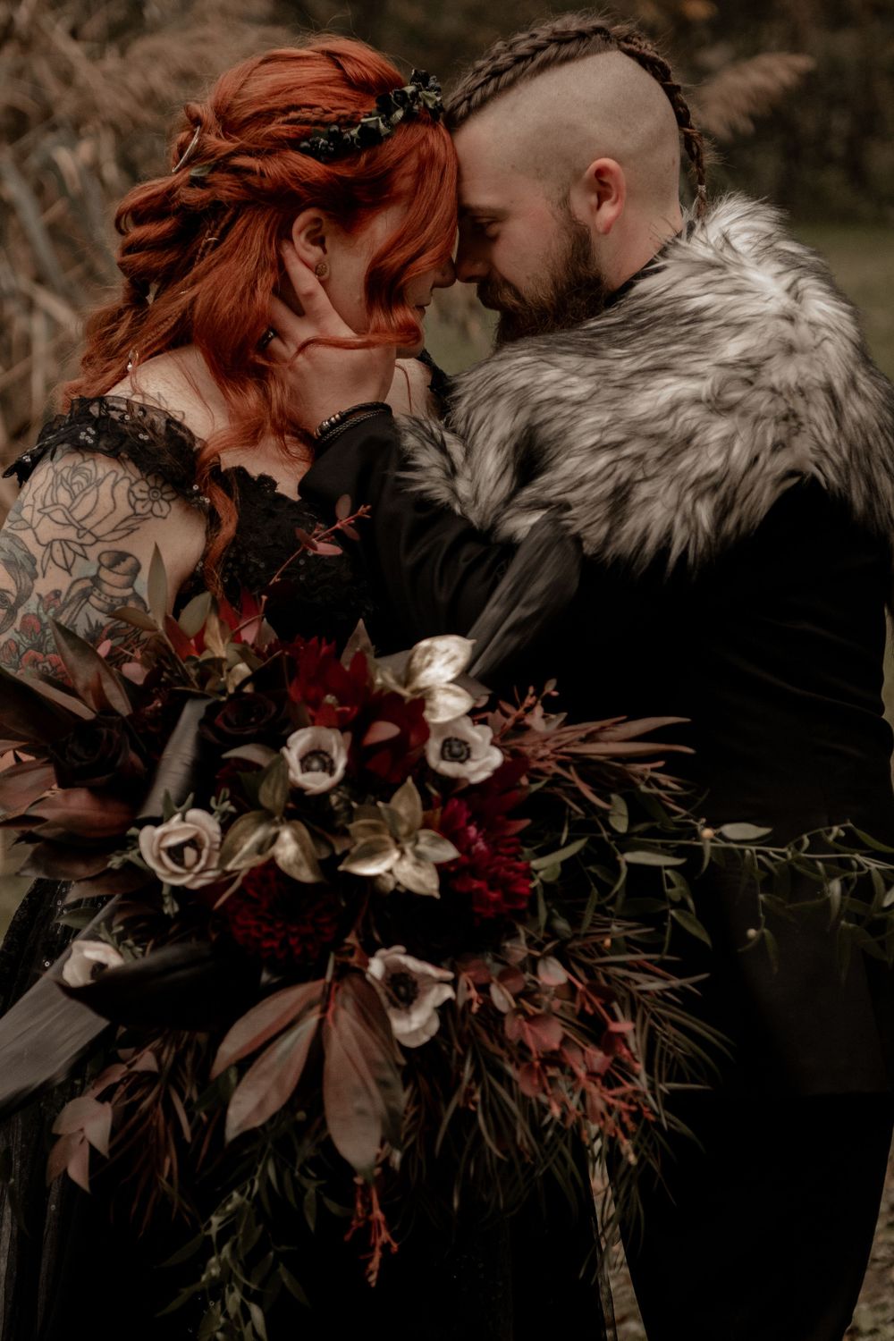 Tattooed redhead bride in black gown holds dark floral bouquet as groom in fur cape touches her face.