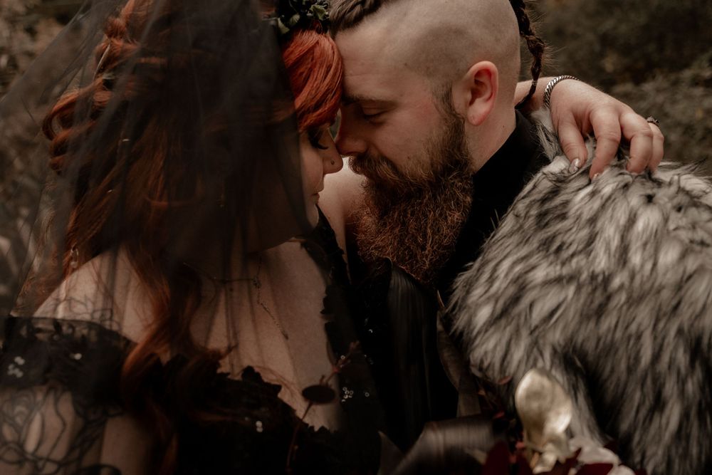 Tattooed redhead bride and bearded groom in fur cape share an intimate moment at their gothic wedding.