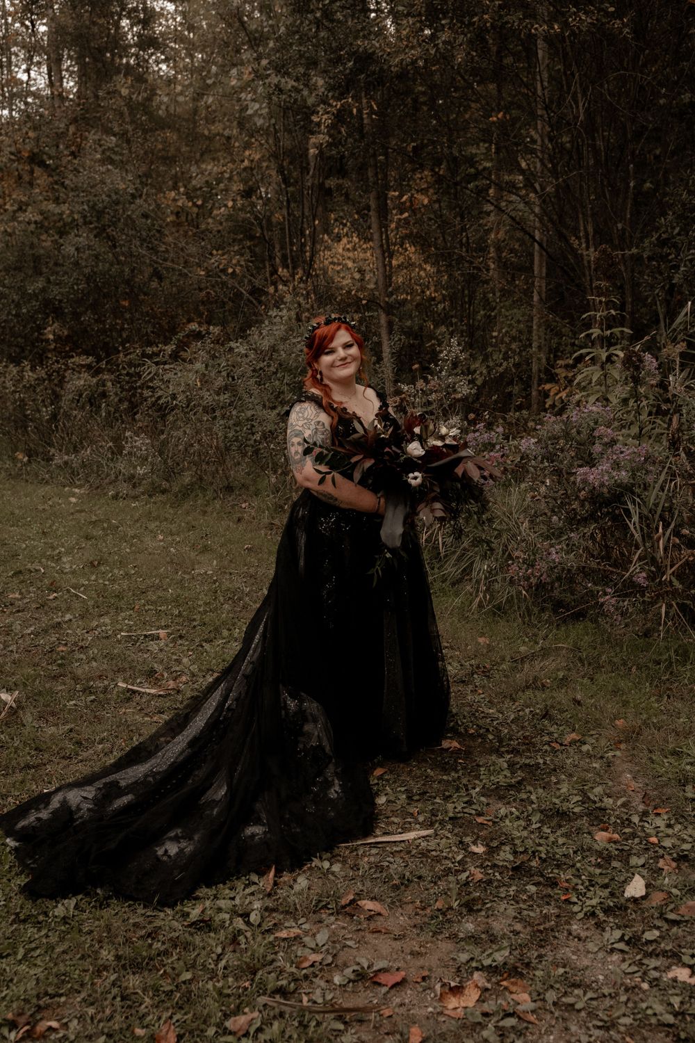 Redhead bride in flowing black lace wedding gown with train holds dark floral bouquet in a forest setting.