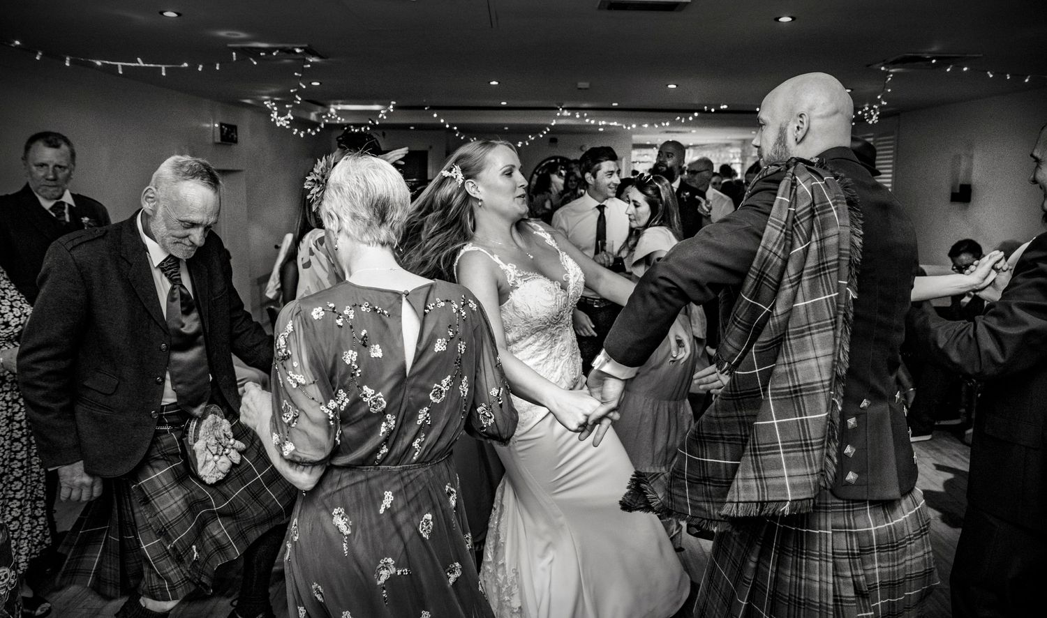 Wedding reception guests dance and celebrate in a dimly lit venue with string lights overhead.