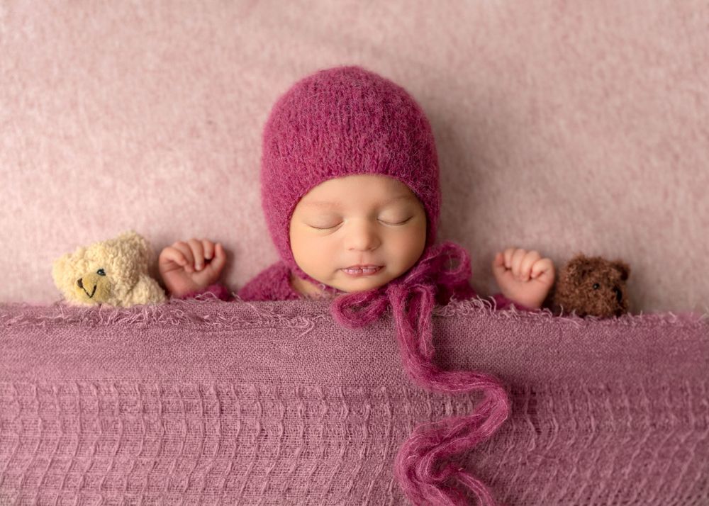 A tiny sleeper wrapped in soft pink fabric with a matching bonnet lies peacefully between two small teddy bears.