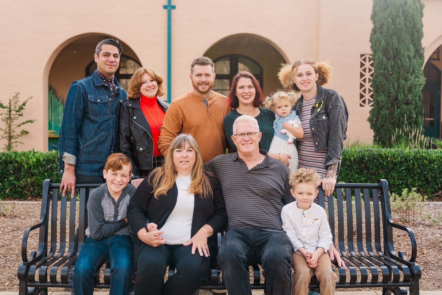 A multi-generational family poses together on benches outside a Mediterranean-style building with arched doorways.