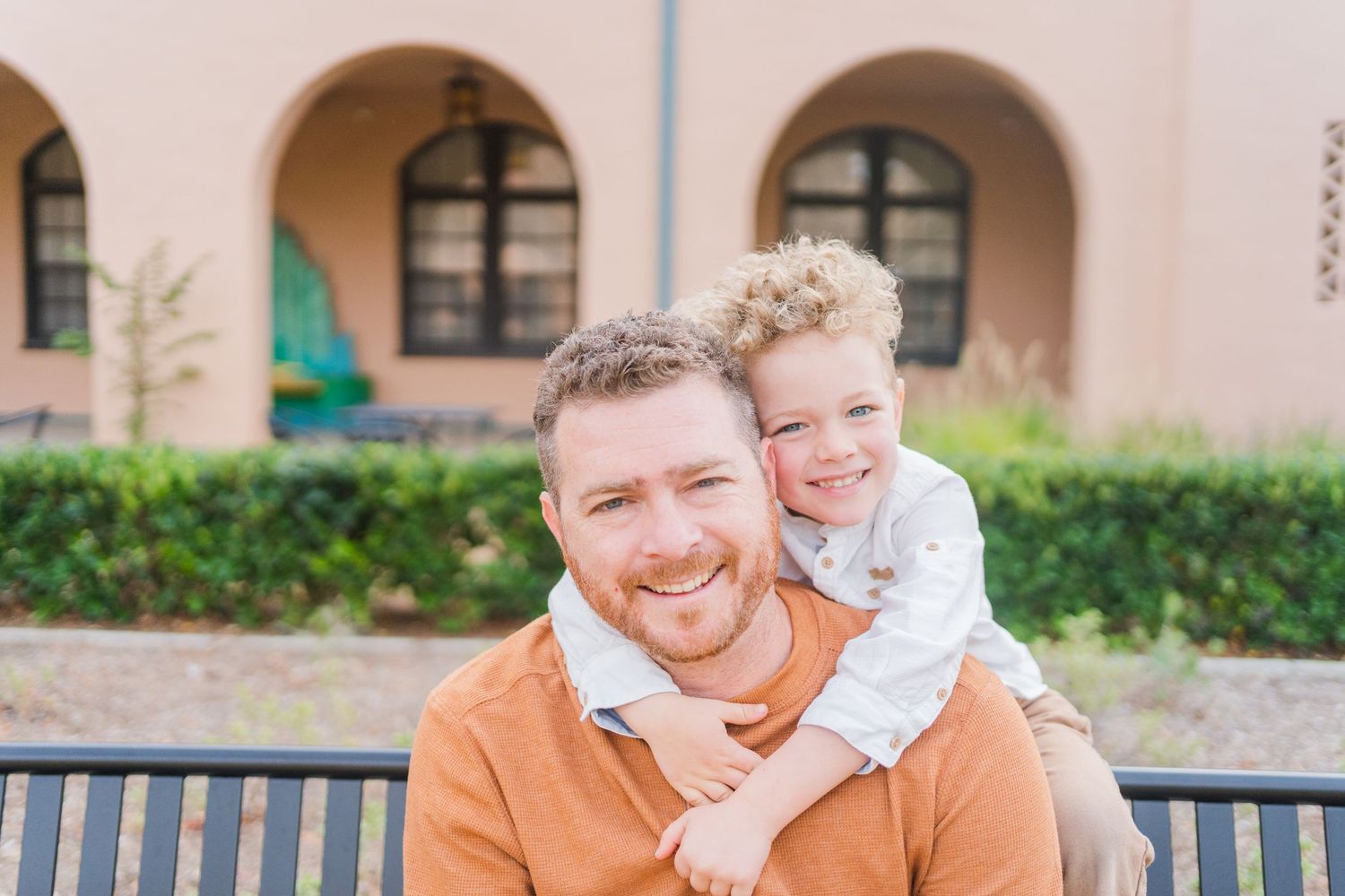 A person and child smile together while sitting on a bench in front of an arched building.