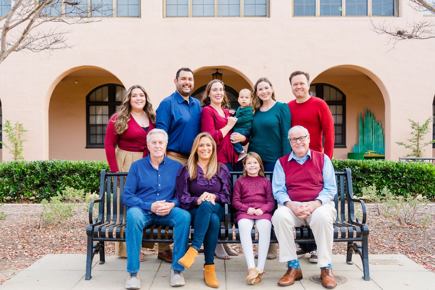 A group of people wearing red, blue and burgundy colored clothing pose together on a black bench outside a building.
