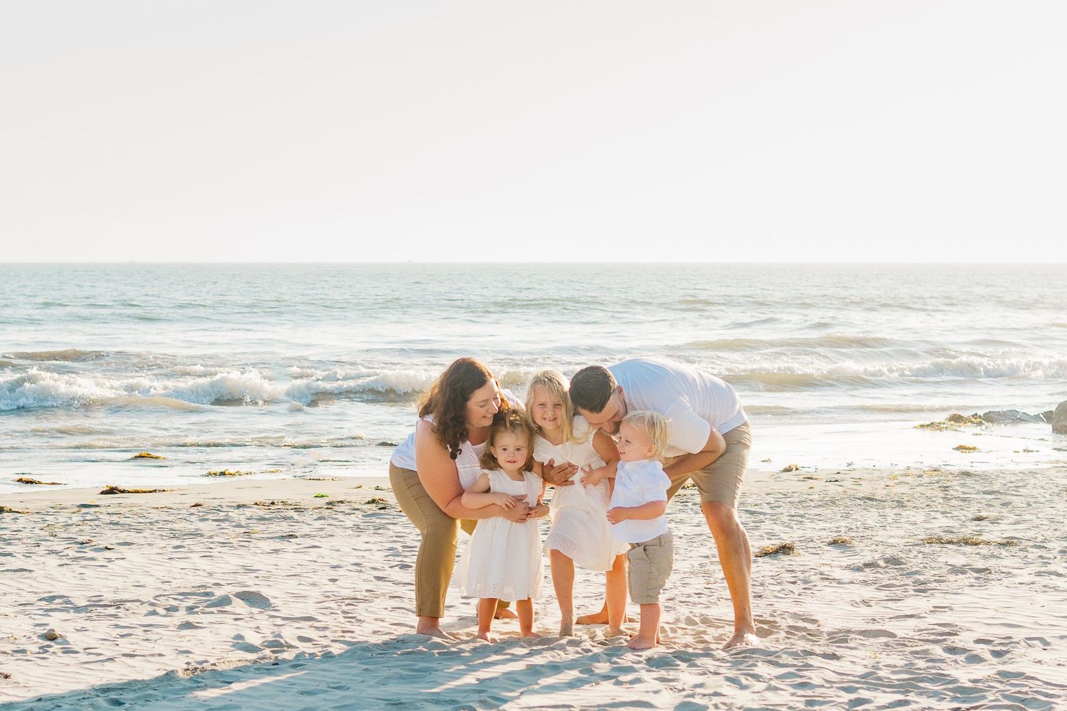 A family dressed in white gathers together looking down at something on a sunny beach with waves in the background.