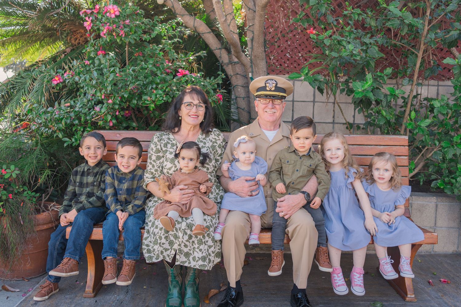 A group of children and adults sit together on a wooden bench in a garden setting with blooming trees and plants.