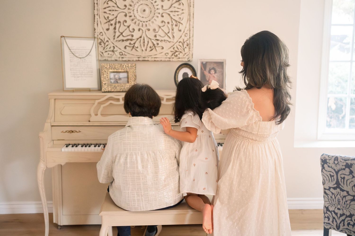 A family sits together at a beige piano in a sunlit room with bohemian decor and wall art.