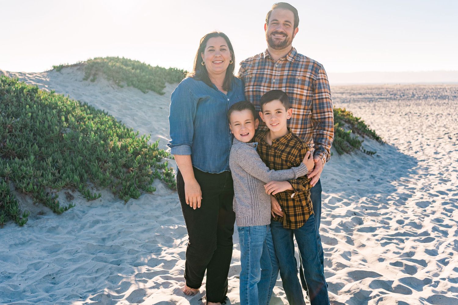 A family of four stands together on a sunny beach with sandy dunes and coastal vegetation in the background.