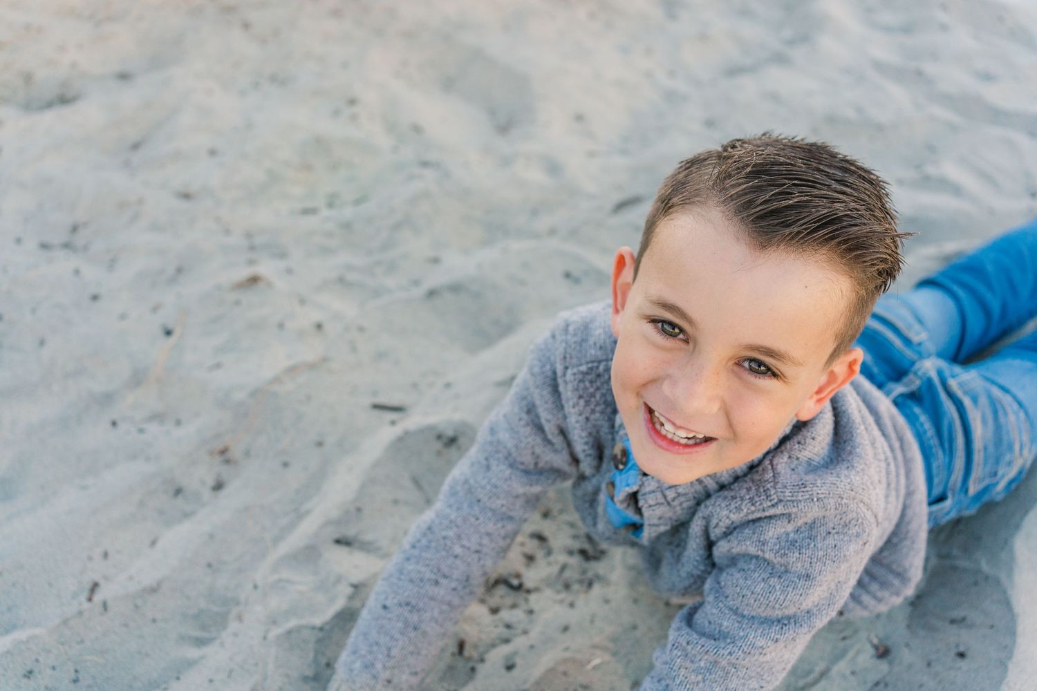 Young child in gray sweater lying on sand at the beach with a bright smile.