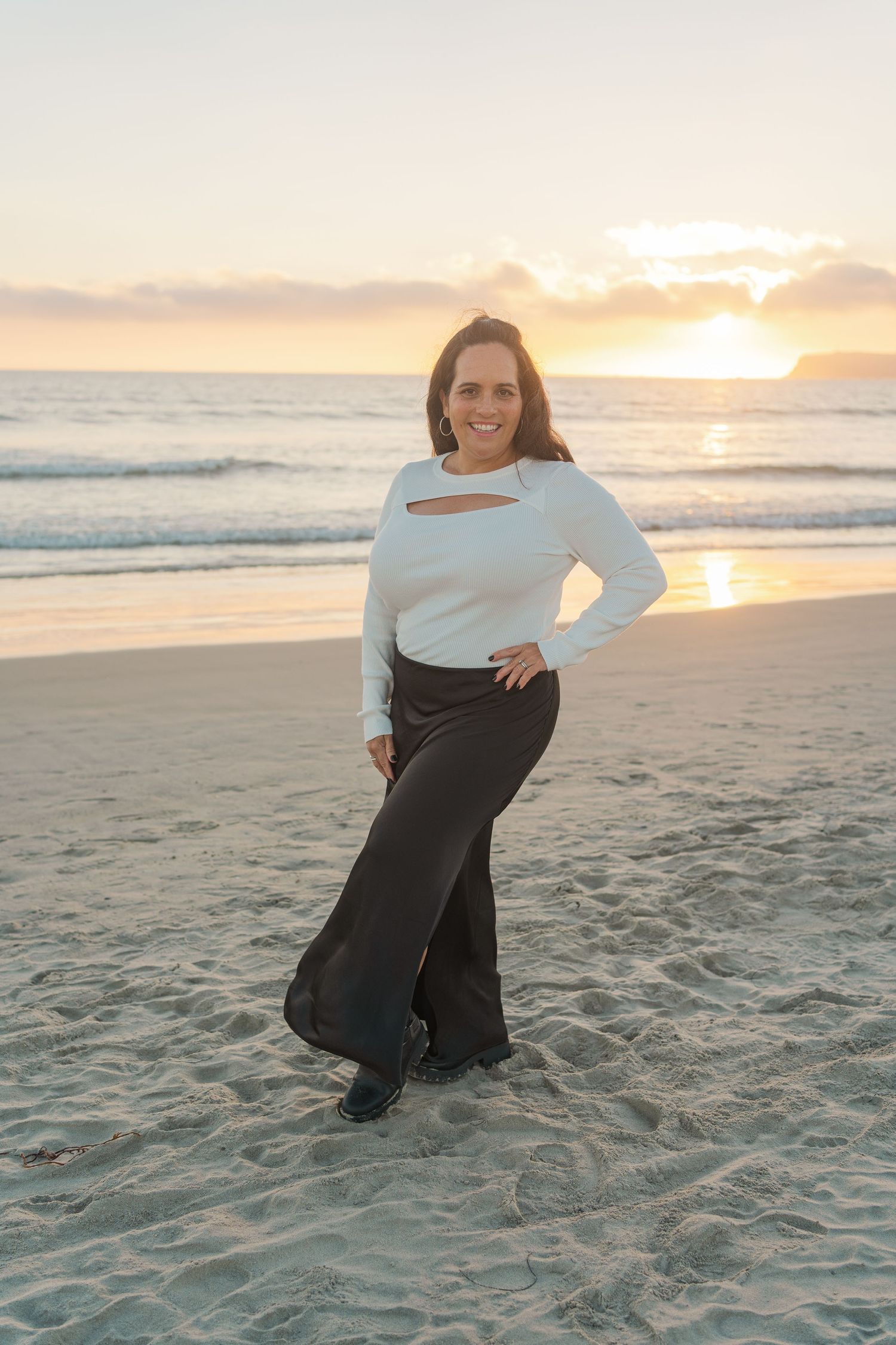 Person in white sweater and black pants poses on beach at sunset with waves and golden sky in background.