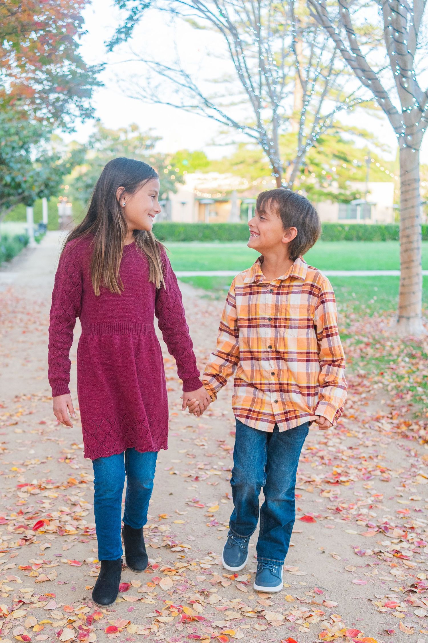 Two children walking together and holding hands on an autumn path with fallen leaves and trees in the background.