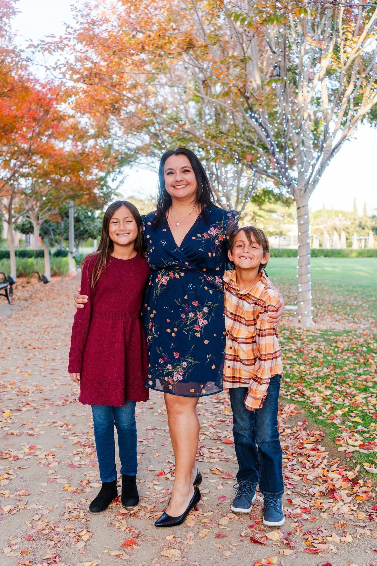 A family poses together on an autumn day with fall foliage and colorful leaves scattered on the ground in a park setting.