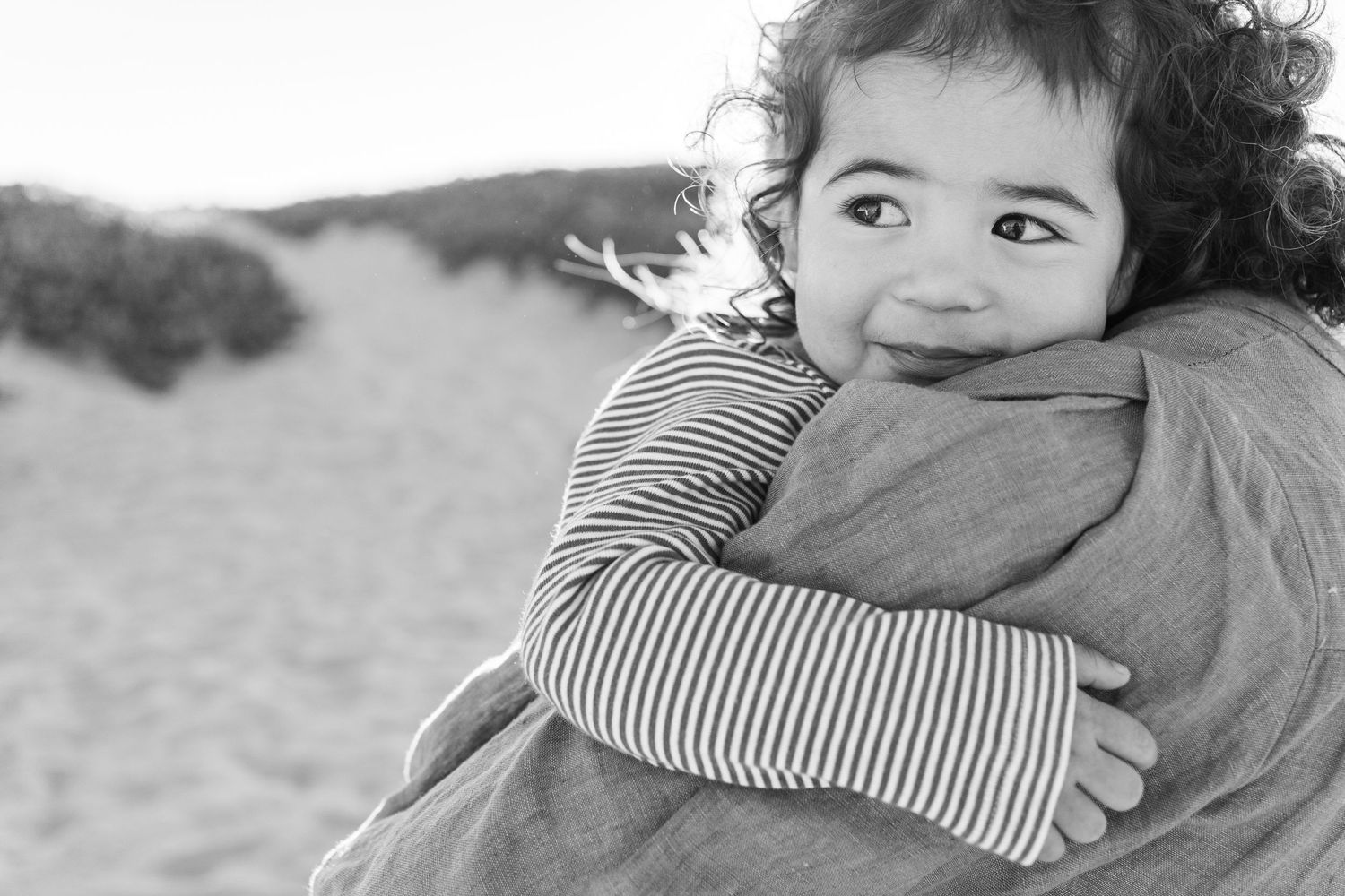 Black and white portrait of a small child in striped sleeves smiling while hugging someone at the beach.