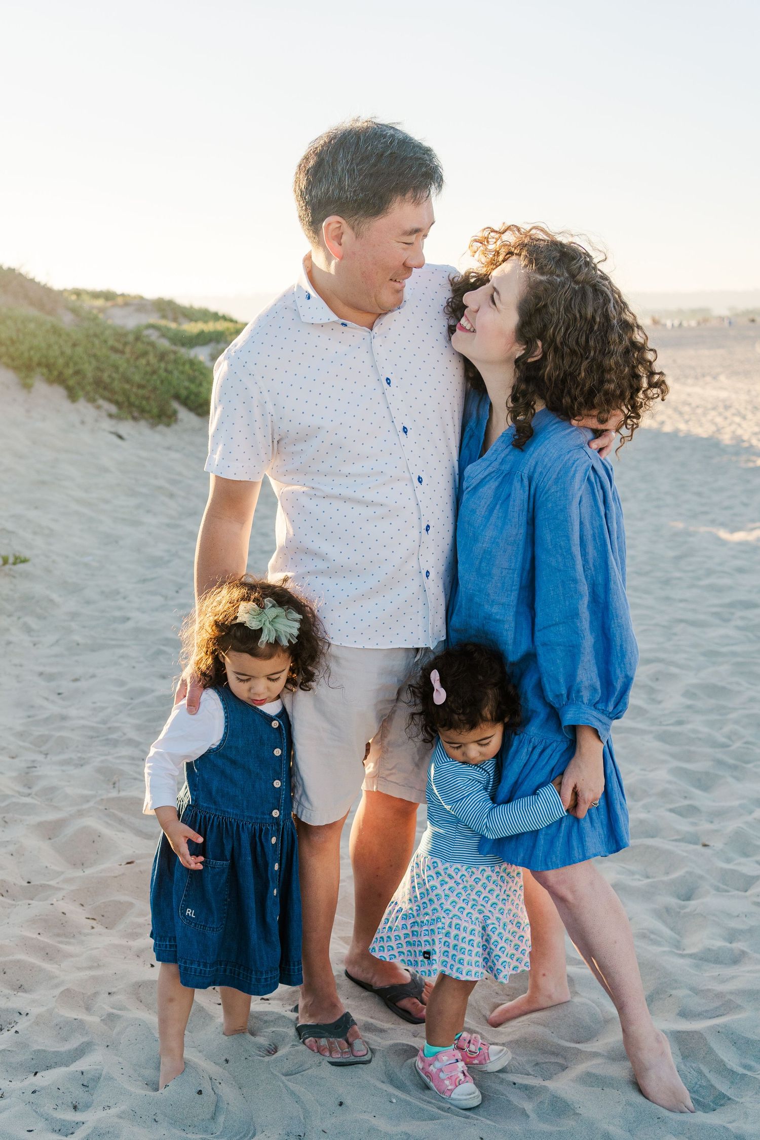 Family wearing coordinated blue and white outfits smiling together on a sandy beach at sunset.