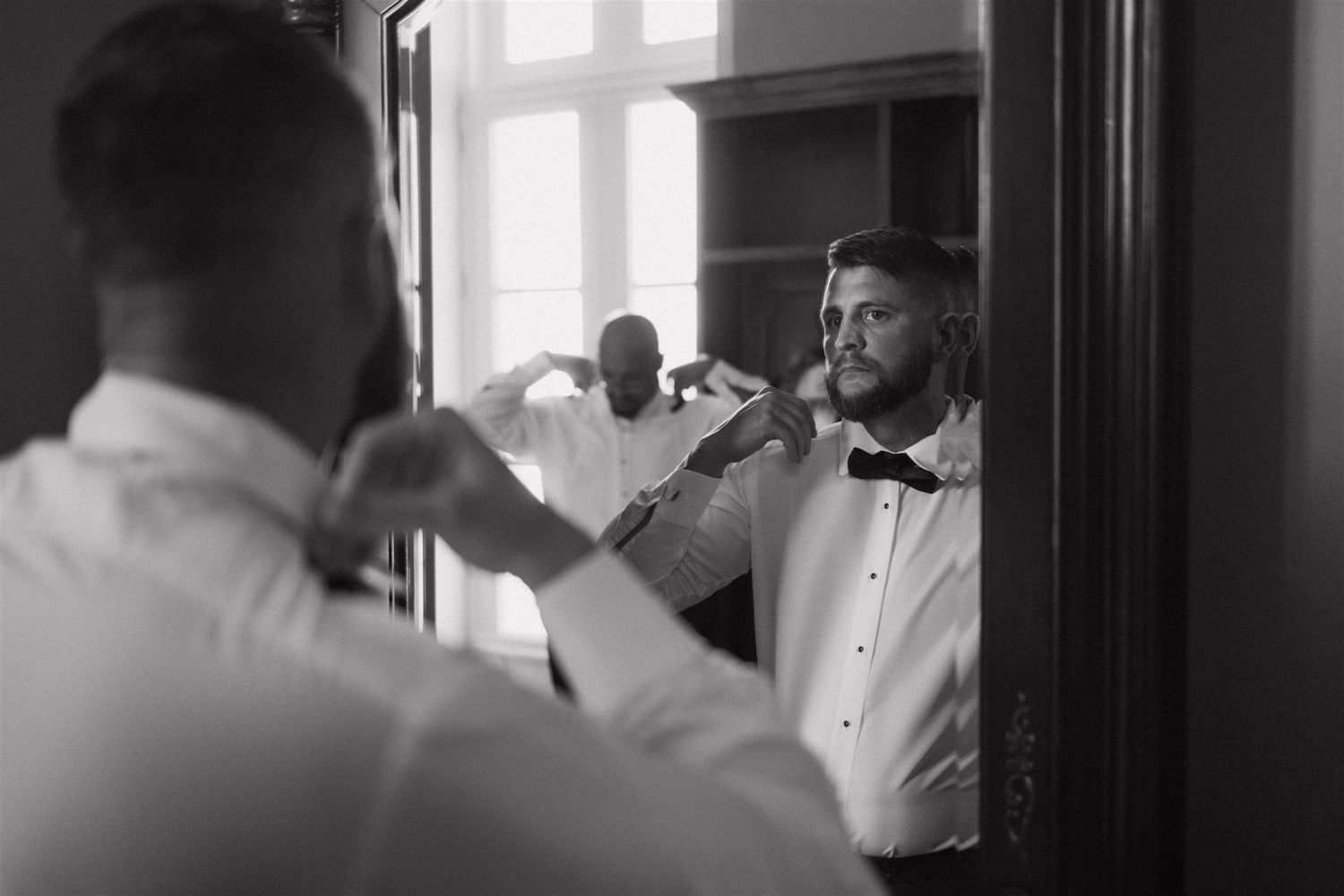 A black and white image shows groomsmen adjusting their bow ties while getting ready in a bright room with windows.