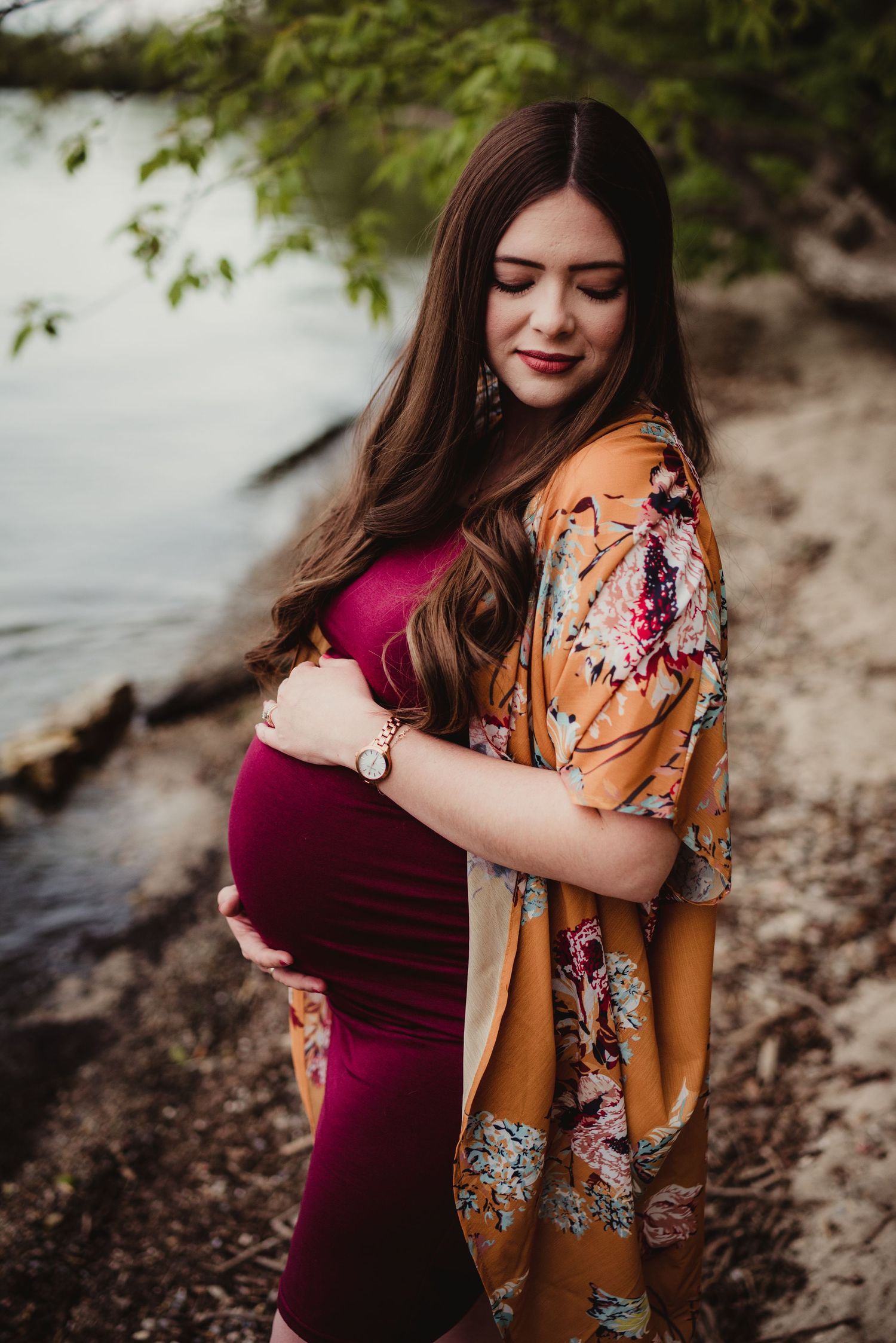 A couple in burgundy and orange floral outfits embrace tenderly by the waterfront during a photo session.