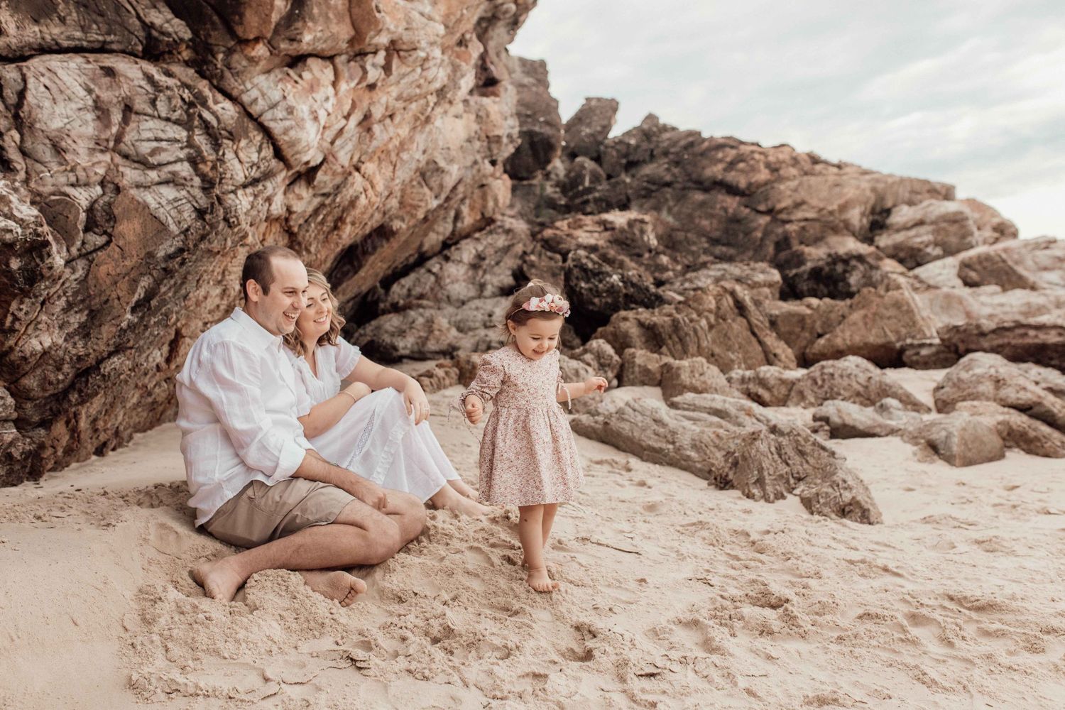 Joyful family moment on a Gold Coast beach, with parents sitting on the sand and smiling as their young daughter plays