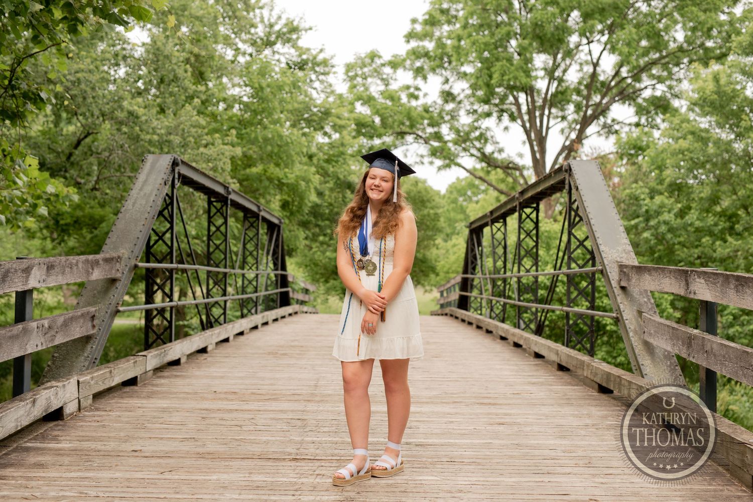 A graduate in white dress poses on a wooden bridge with iron railings.