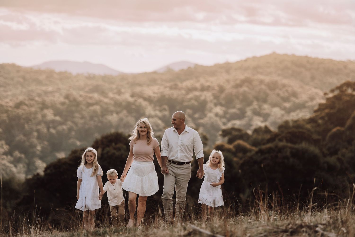 A family stands together on a hilltop during golden hour, with mountains and trees in the background.