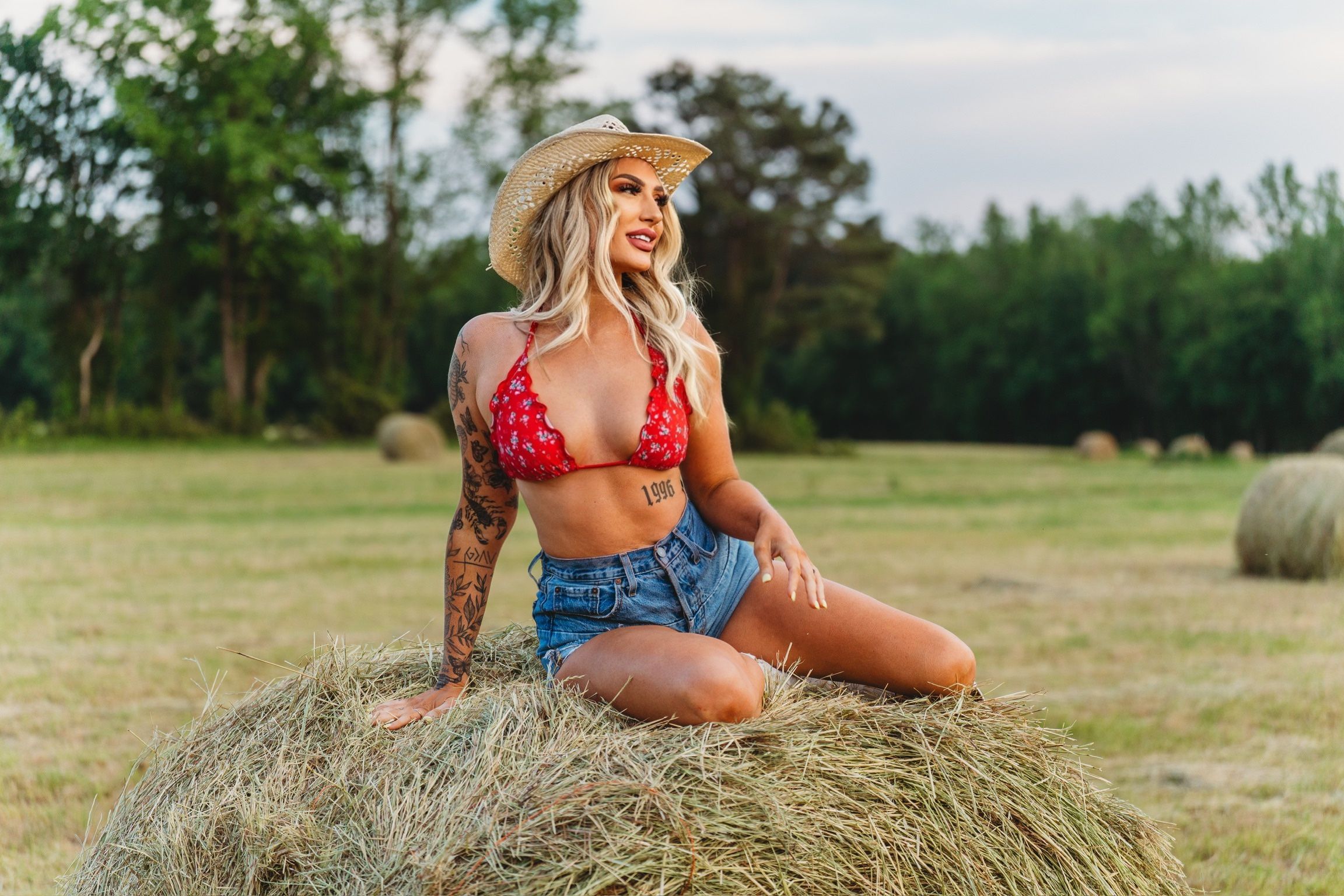 Woman in red bikini top and denim shorts sitting on hay bale in field wearing cowboy hat.