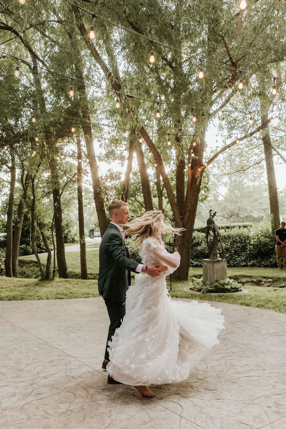 Alpine Utah couple dances and twirls in wooded area with streaming sunlight and hanging lights.
